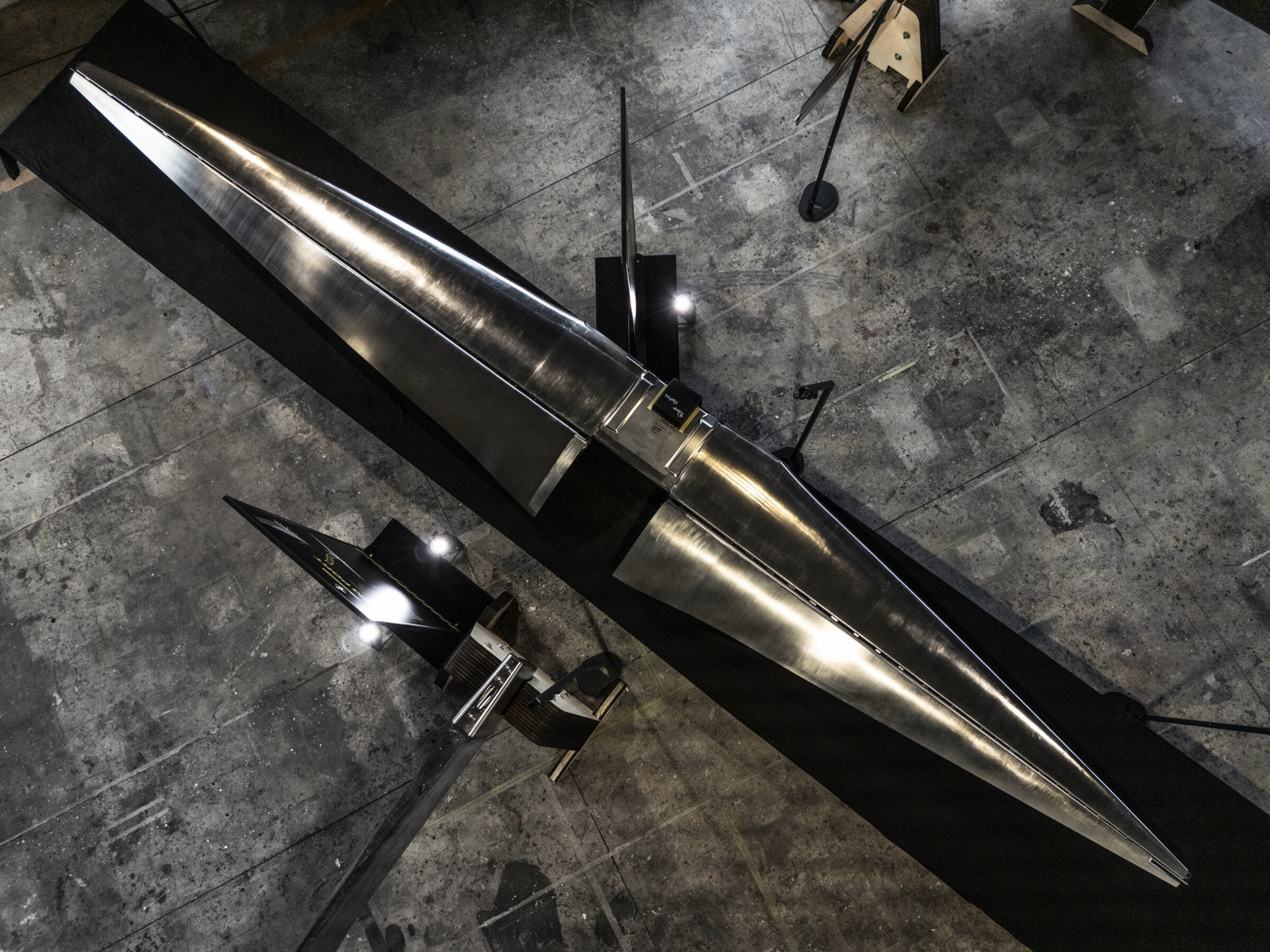 A long polished appendage seen from above, lying on a black stand on the shipyard floor.